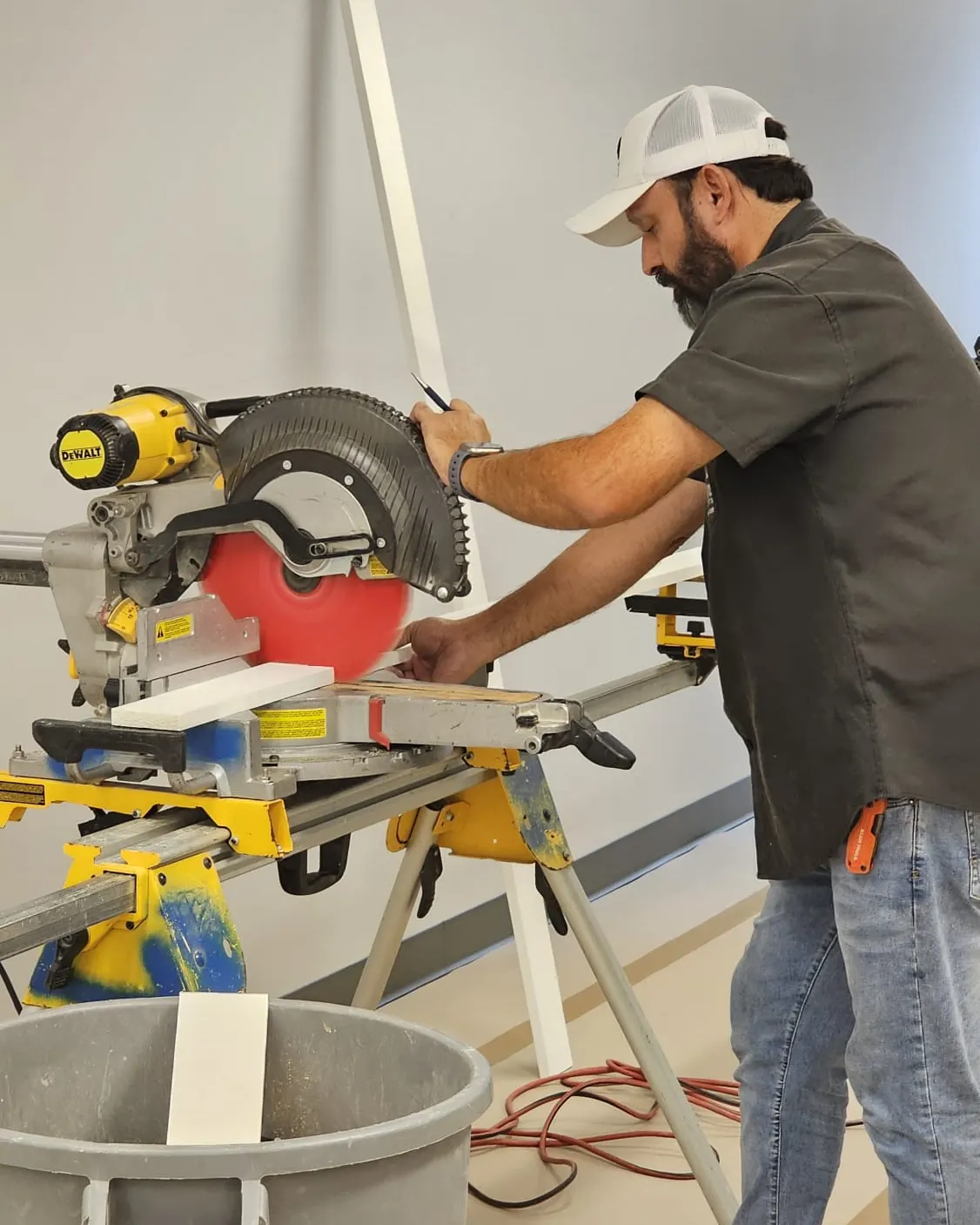 Carpenter cutting white trim stock on a miter saw with a waste bin in front